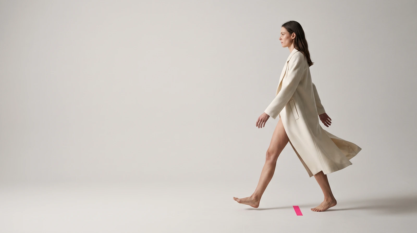 A model caught mid-stride across a warm grey studio sweep, rear heel lifted and legs scissored apart, the left half of the frame empty paper, a short strip of magenta tape stuck to the floor at the spot she has just left